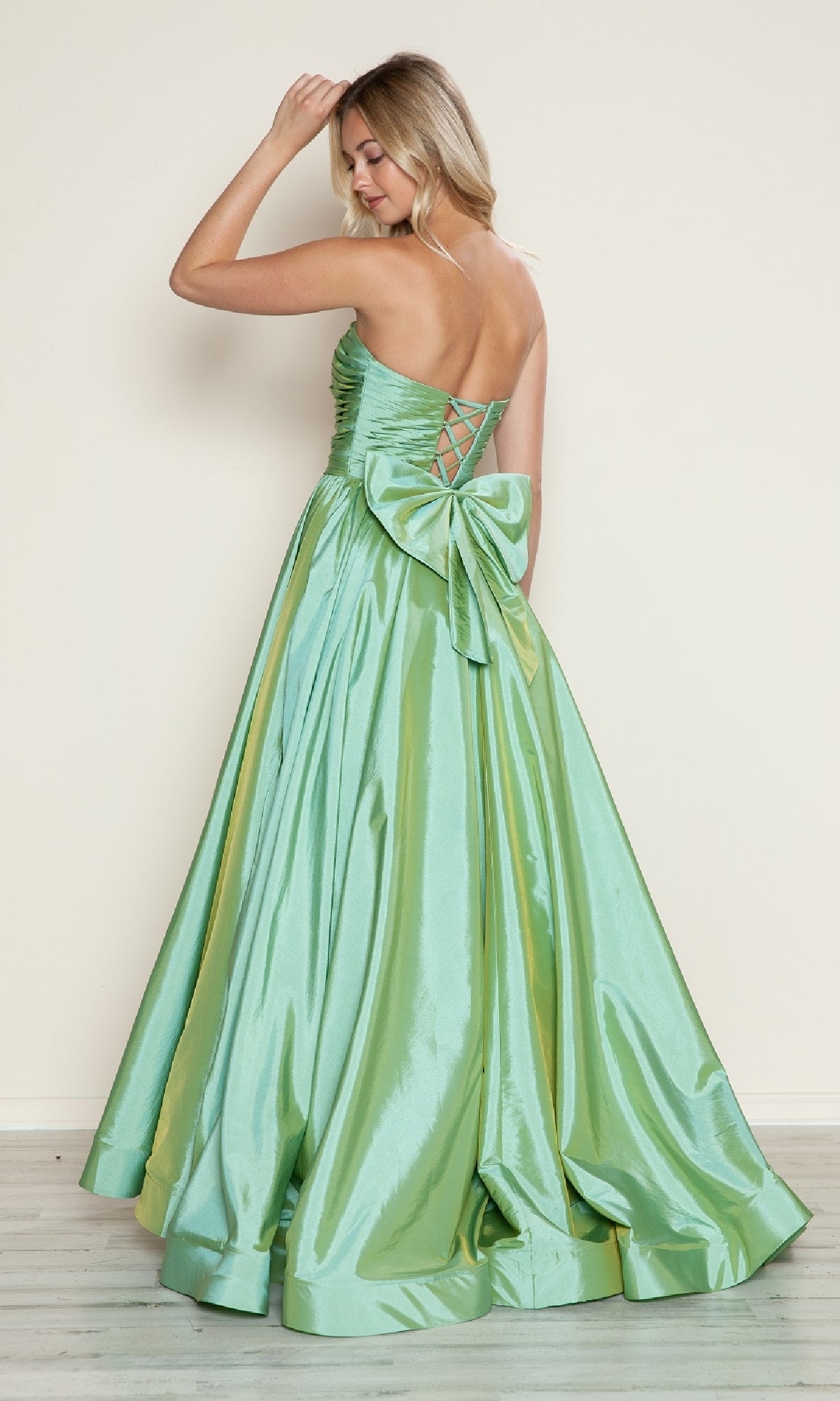 Wearing the Poly USA 9754 long prom dress, a woman stands facing away, touching her hair. The shiny light green taffeta gown features a strapless ruched bodice, adjustable lace-up back, and a large accent bow enhancing her elegant style.